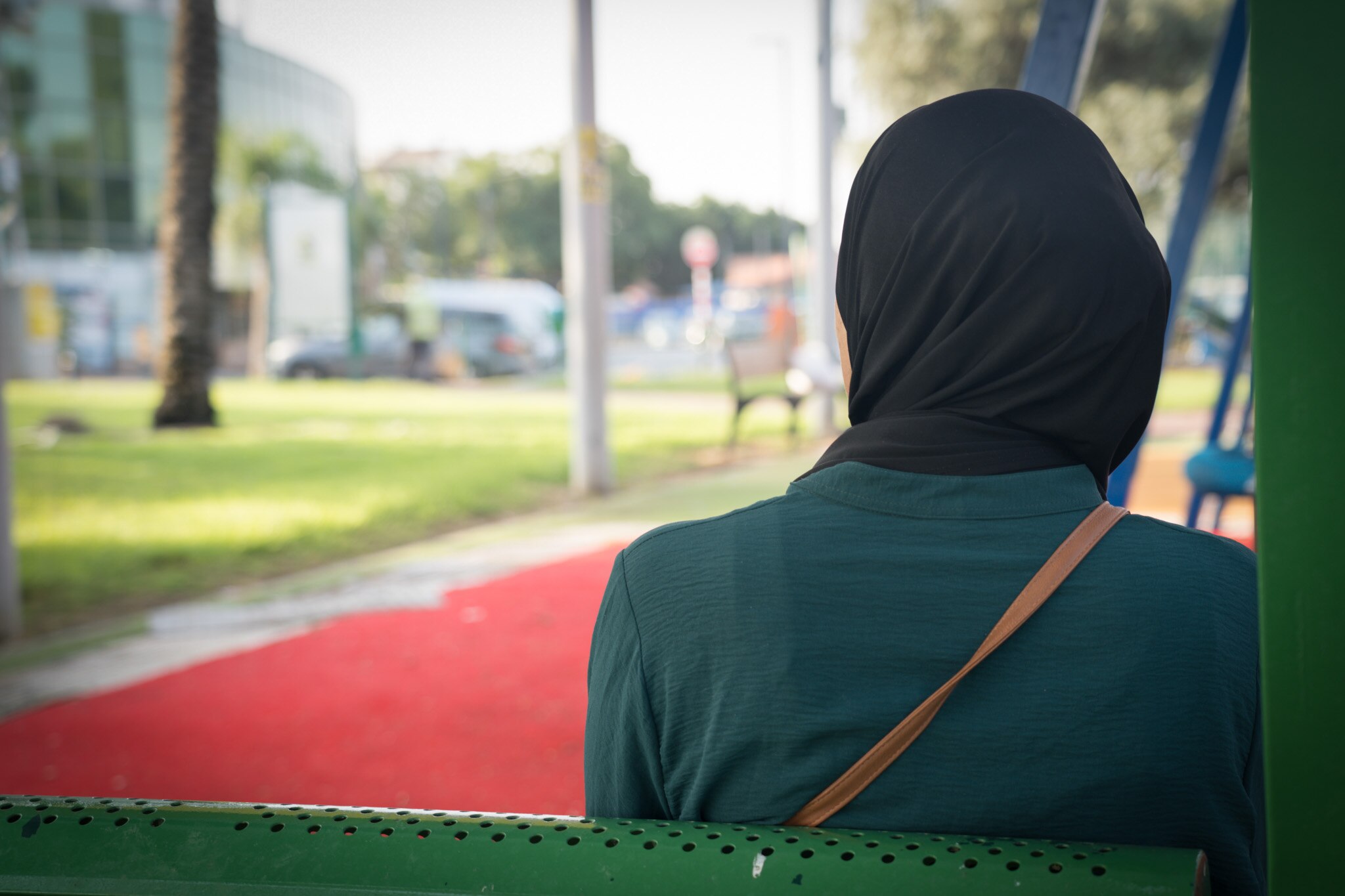 A close up of the back of a woman's head as she sits on a bench. A park with a swing is in the background.