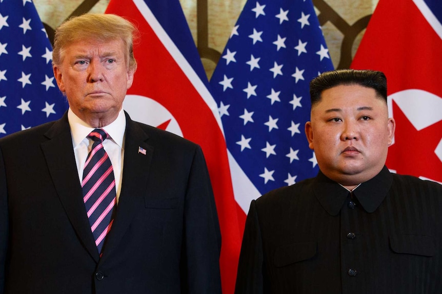 Donald Trump and Kim Jong-un stare stony-faced while standing in front of North Korean and United States flags.