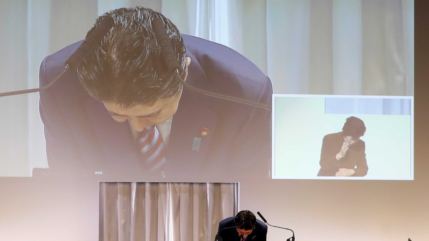 Shinzo Abe is seen bowing as he stands on a podium, behind him we see the projection of his image and a woman bowing.