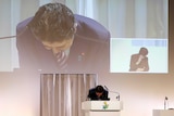 Shinzo Abe is seen bowing as he stands on a podium, behind him we see the projection of his image and a woman bowing.