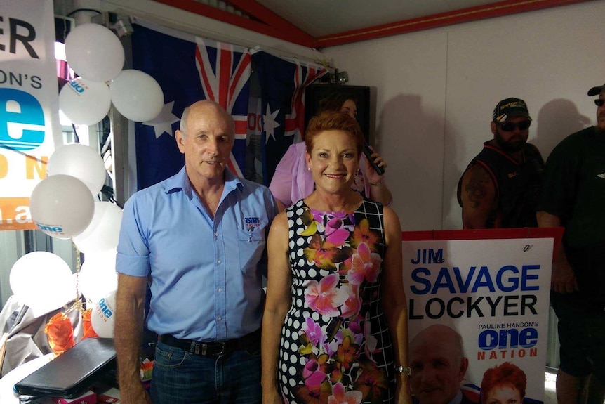 A balding man in a blue shirt stands beside a grinning woman in a floral dress in front of an Australian flag.