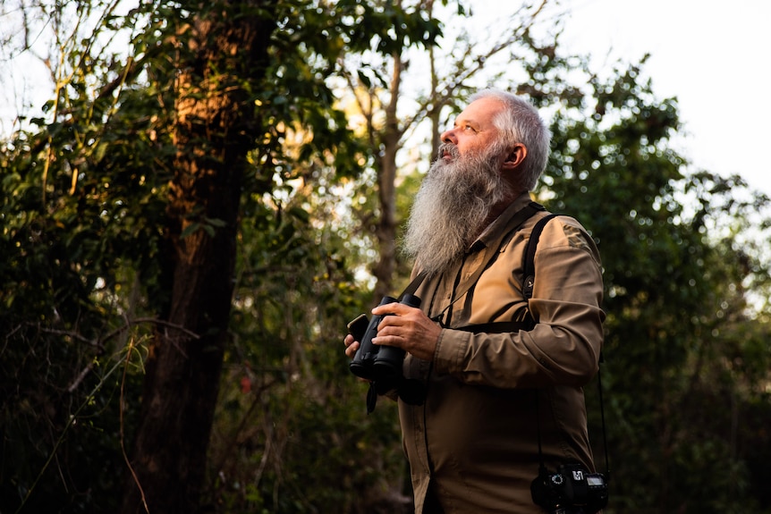 A man with a beard and binoculars looks to the trees. 