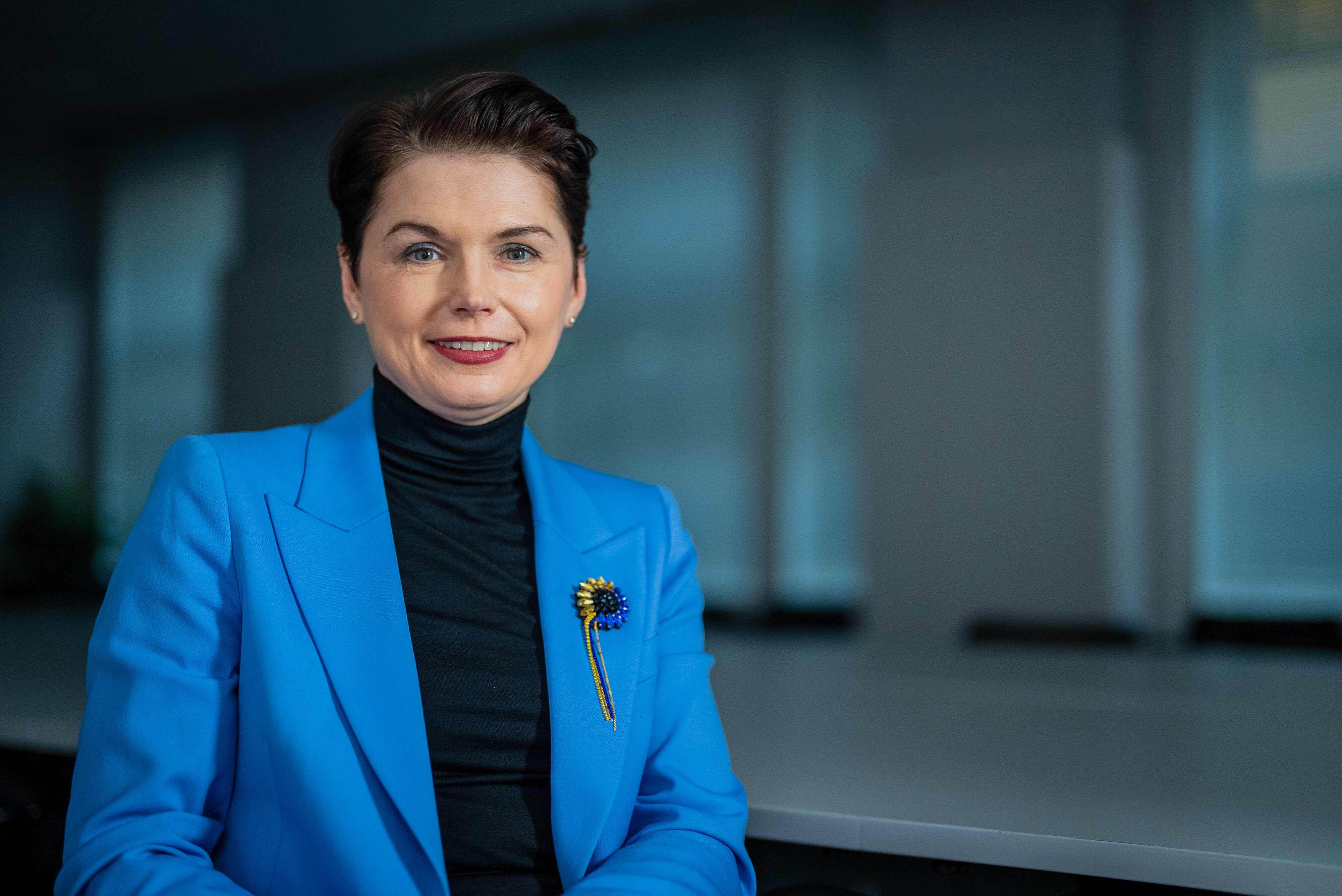 A woman with short brown hair wears a blue jacket smiling at the camera