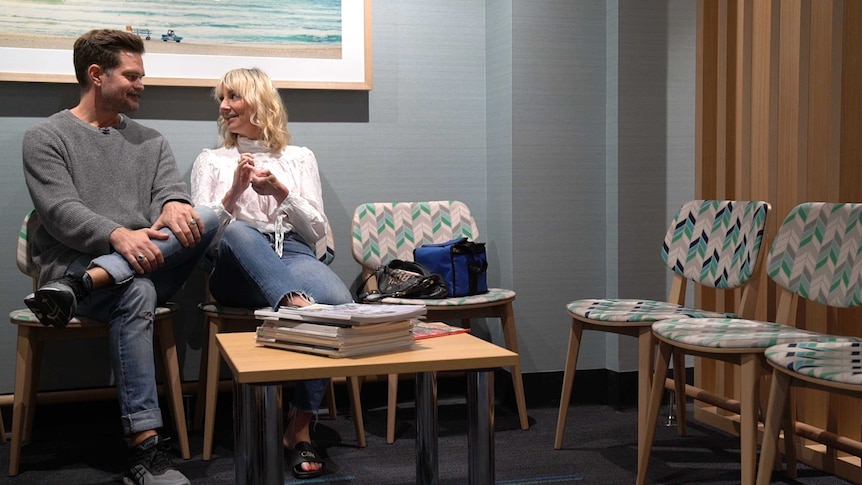 A man and a woman sit beside each other in a hospital waiting room with a coffee table in front of them, ahead of an IUI attempt