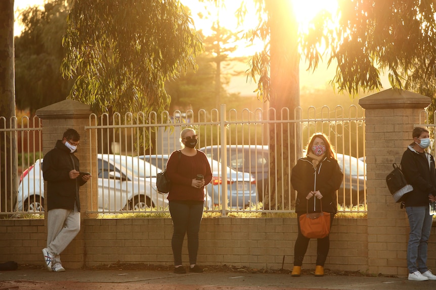 Morning sunshine flares over a line of four people waiting on a footpath wearing face masks. 