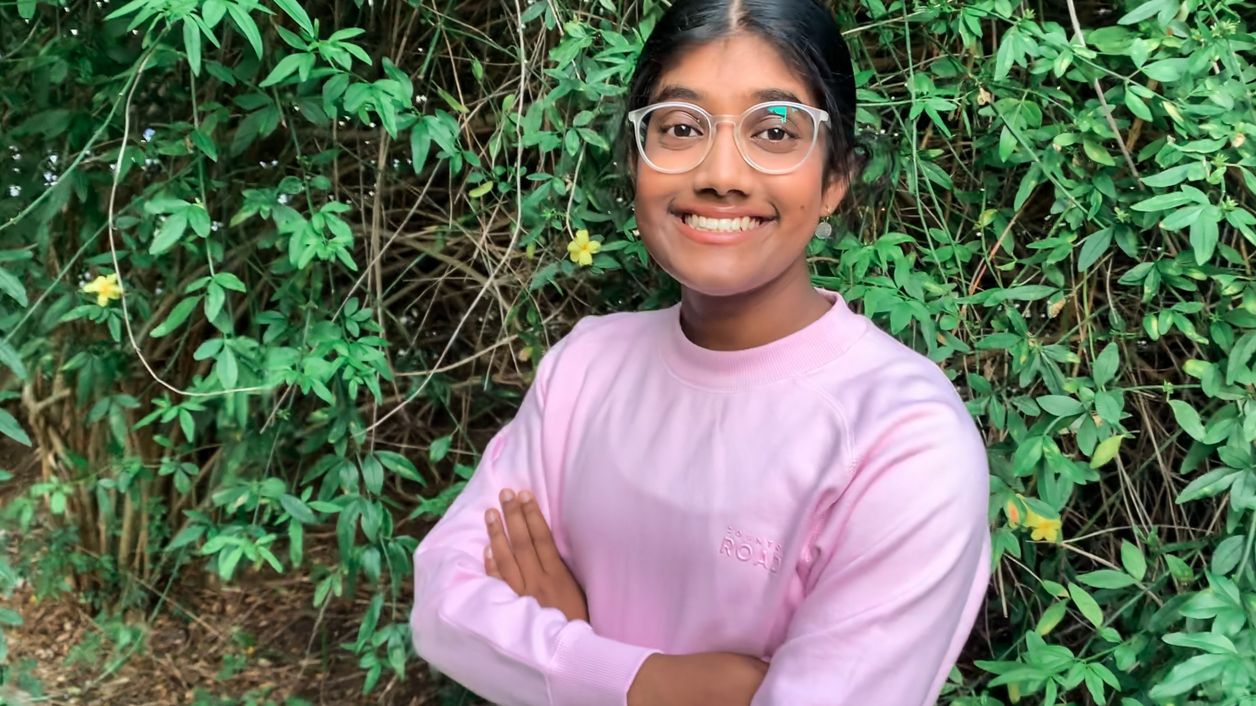 a young woman stands arms crossed in front of a green hedge smiling