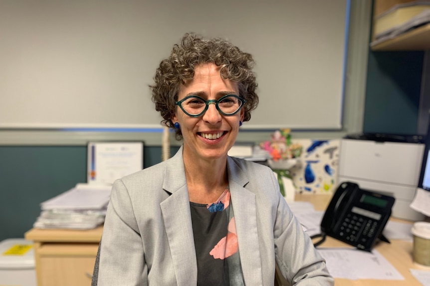 A woman with short curly grey hair, glasses sits in her GP consultation room.