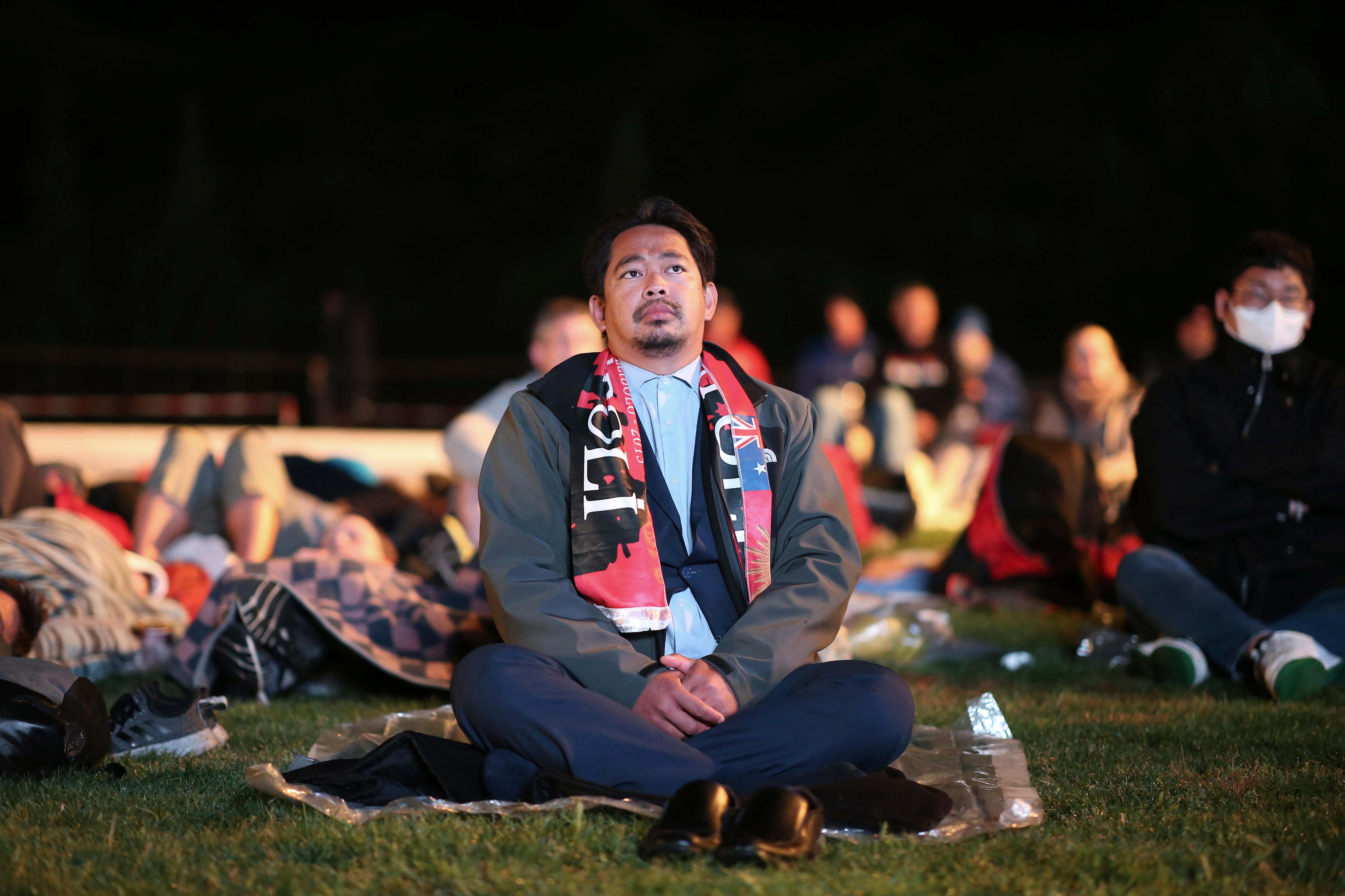 A man with a solemn expression sits on a space blanket on a lawn among a crowd with a black sky above him