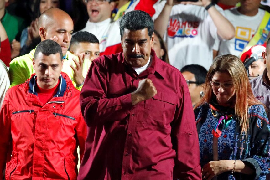 Man with dark hair and moustache wearing red jacket holds fist over heart in front of crowd of people