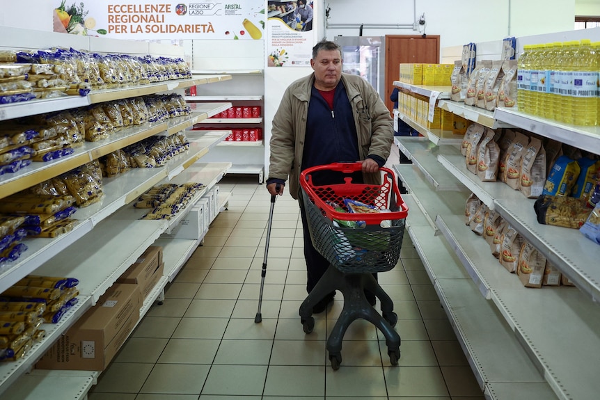 Un hombre camina por el pasillo de un supermercado con un carrito de compras y un bastón.