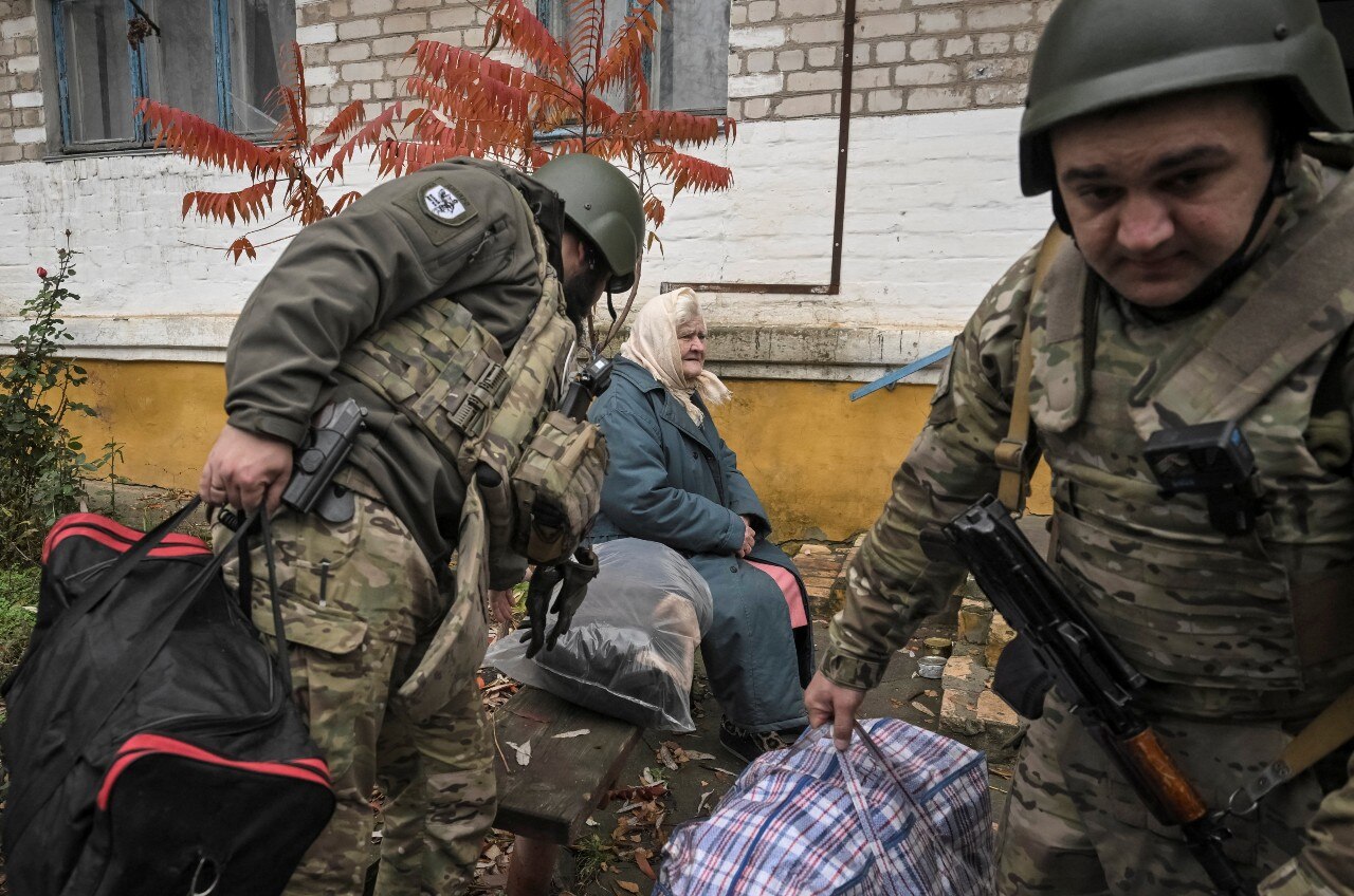 Two men in military uniforms help an elderly woman move