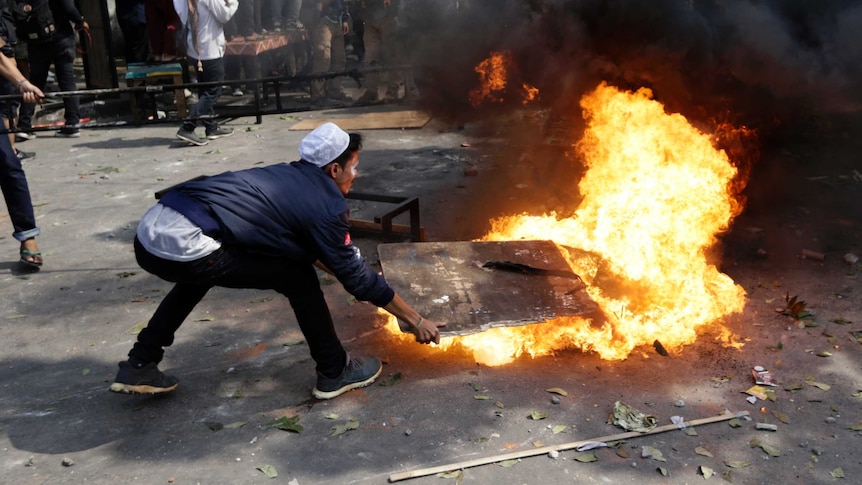 A man squats and puts a large sign over a small but raging fire. Black smoke billows. People congregate in the background.