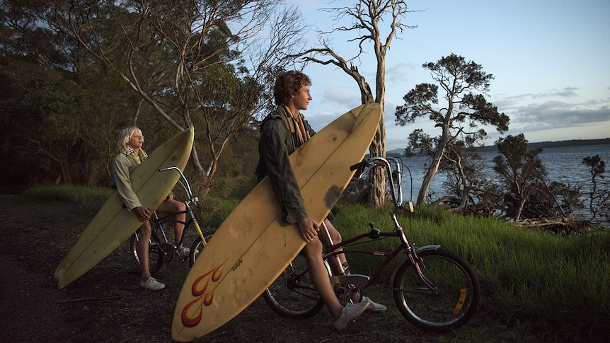 Actors Ben Spencer and Samson Coulter sitting on bicycles, holding surf boards and looking out towards the ocean at sunset.