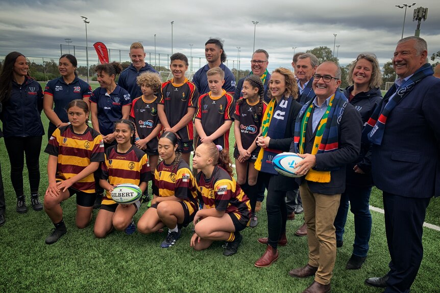 Politicians pose for a photo with Rugby Victoria representatives and a group of rugby players of different ages.