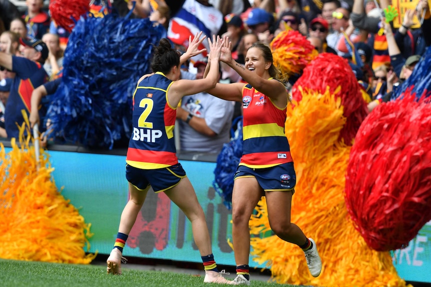 An AFLW player high-fives her teammate after a goal as fans behind her wave their flags.