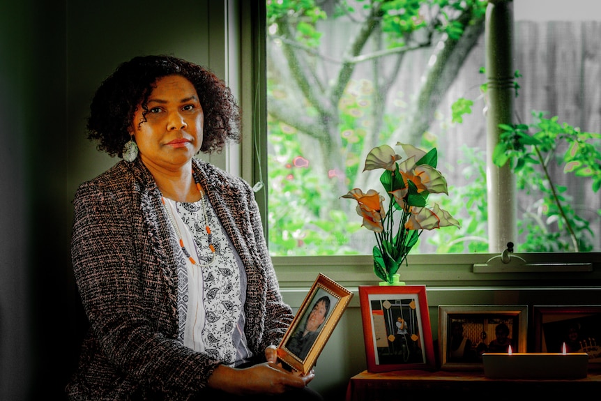 A woman stands by a window holding a framed photo of a man.