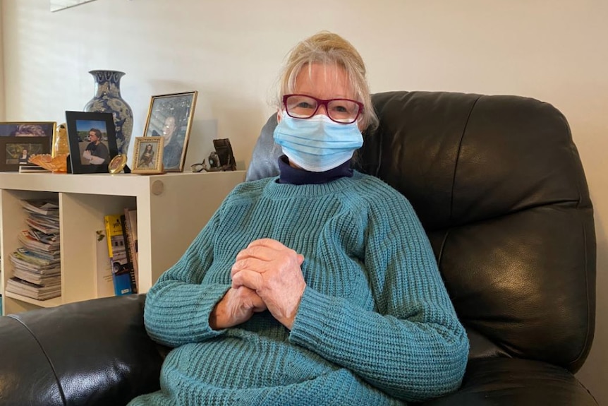 A woman sitting in an arm chair wearing a face mask clasping her hands.