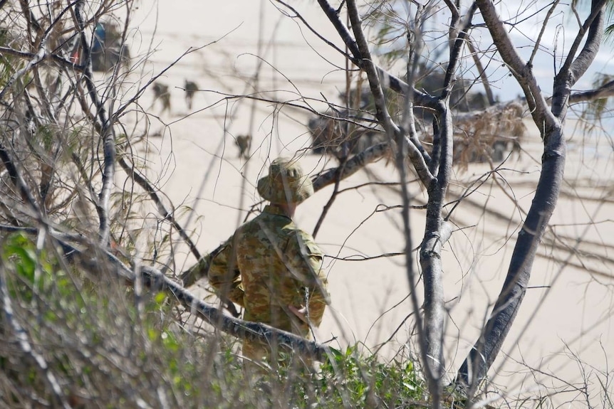 A man in army uniform stands on a hill in the bushes and watches out over a beach where military vehicles are parked.