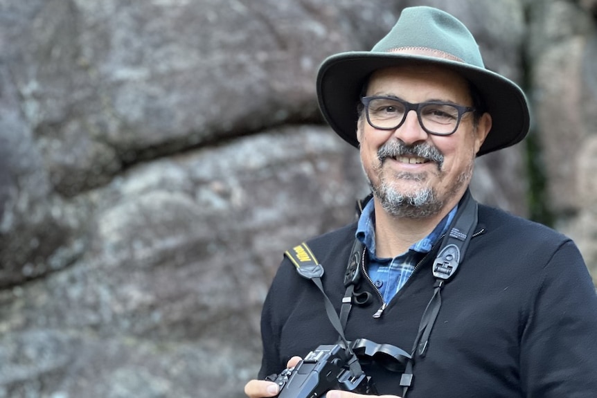 Sean Dooley stands near large rocks with binoculars around his neck and a camera in his hands
