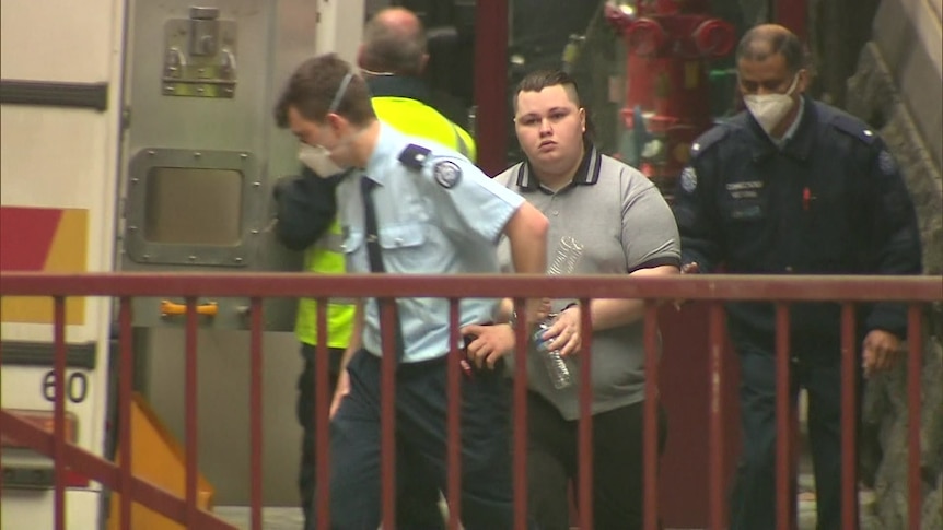 A hand-cuffed man wearing a grey polo shirt surrounded by custody officers.
