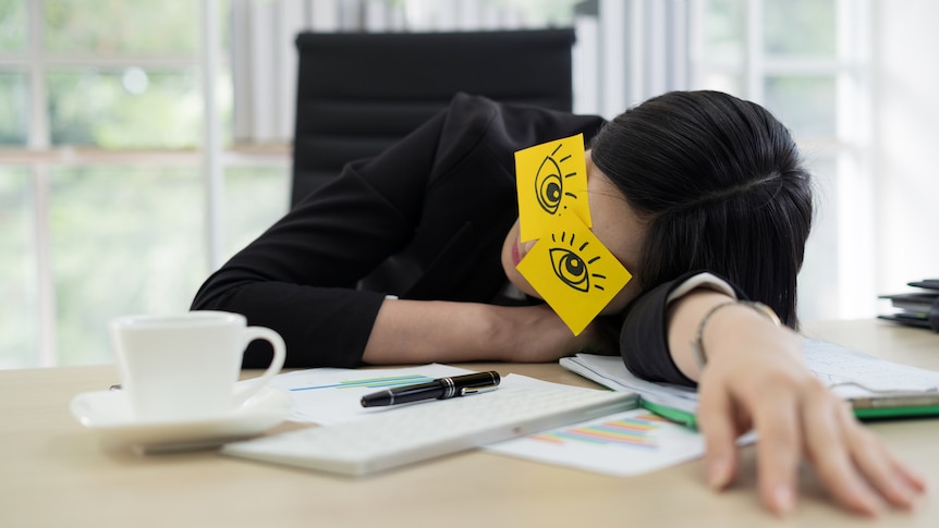 A woman slumps at her desk sleeping, with post it notes on her eyes with cartoon eyes drawn on