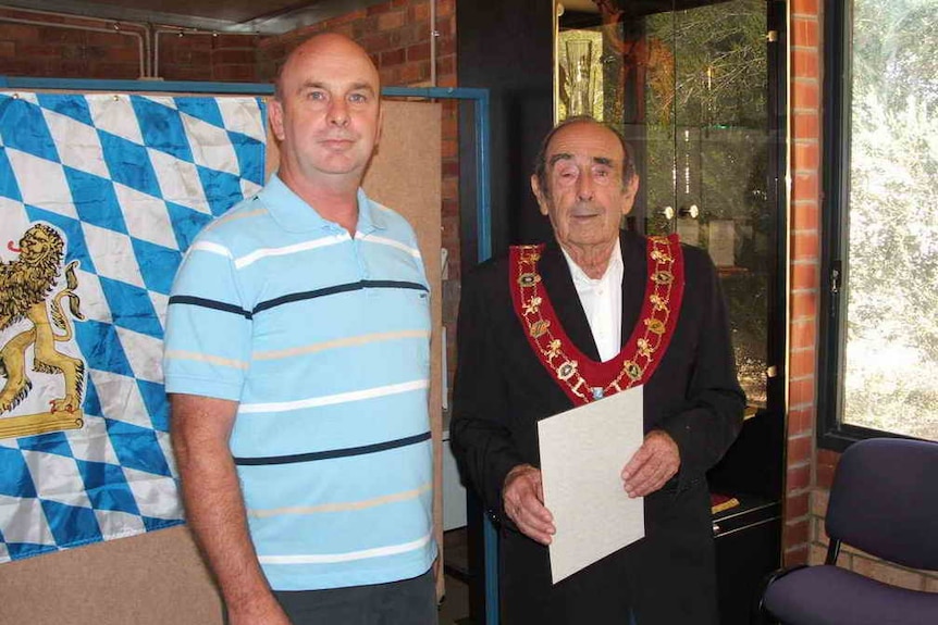 A middle aged man stands next to an older man wearing a ceremonial garland.