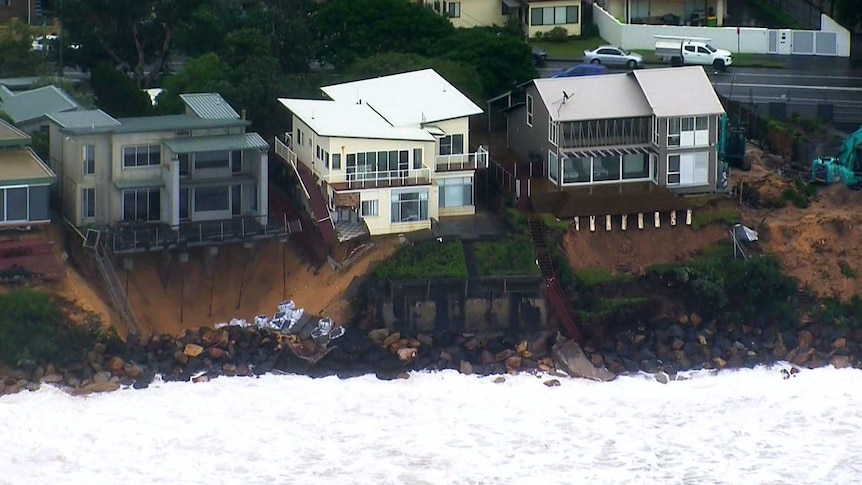 Waterfront houses with exposed foundations are surrounded by landslip as waves crash below.