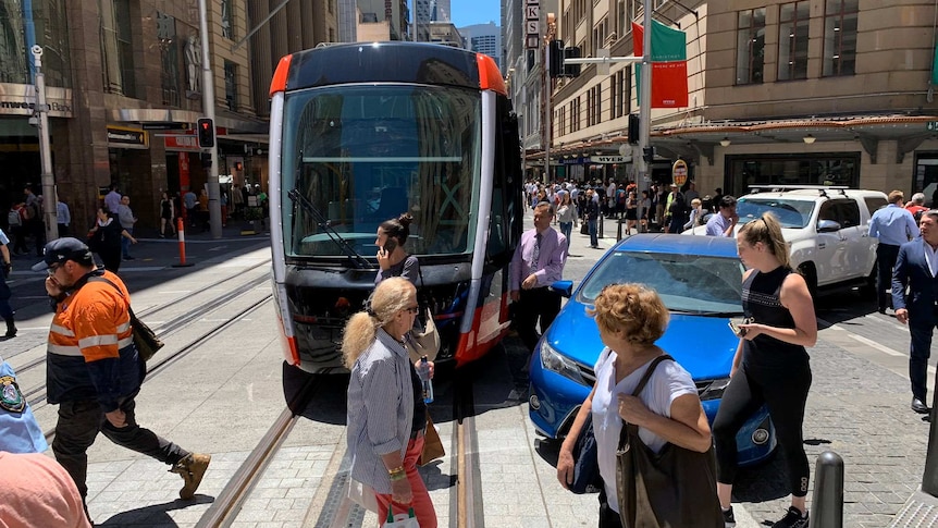 A light rail tram stuck in the middle of the road with cars and people around it.