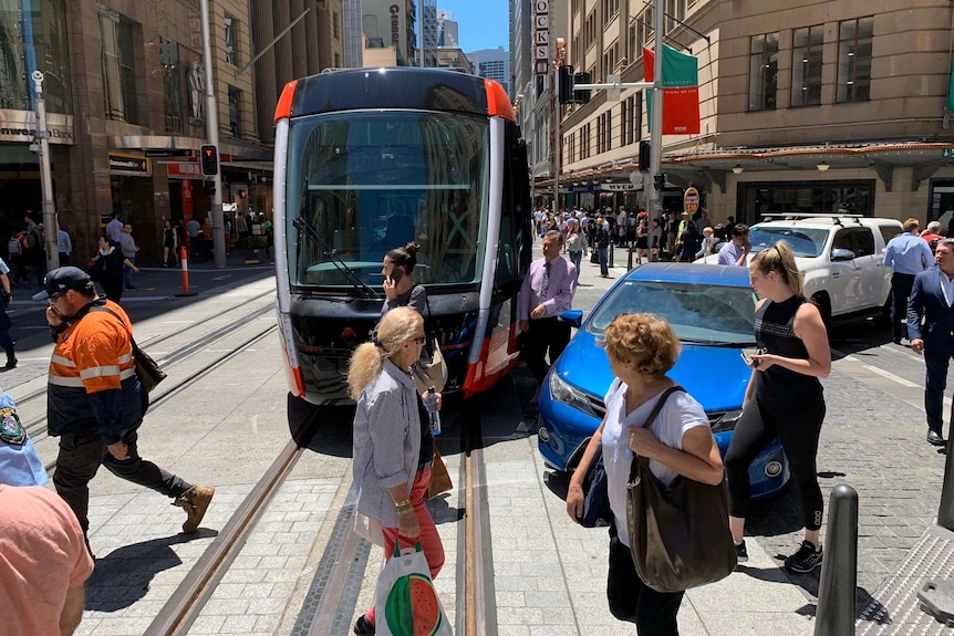 A light rail tram stuck in the middle of the road with cars and people around it.