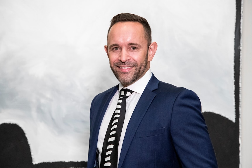 A brown haired man with a beard in a suit looks at the camera. 