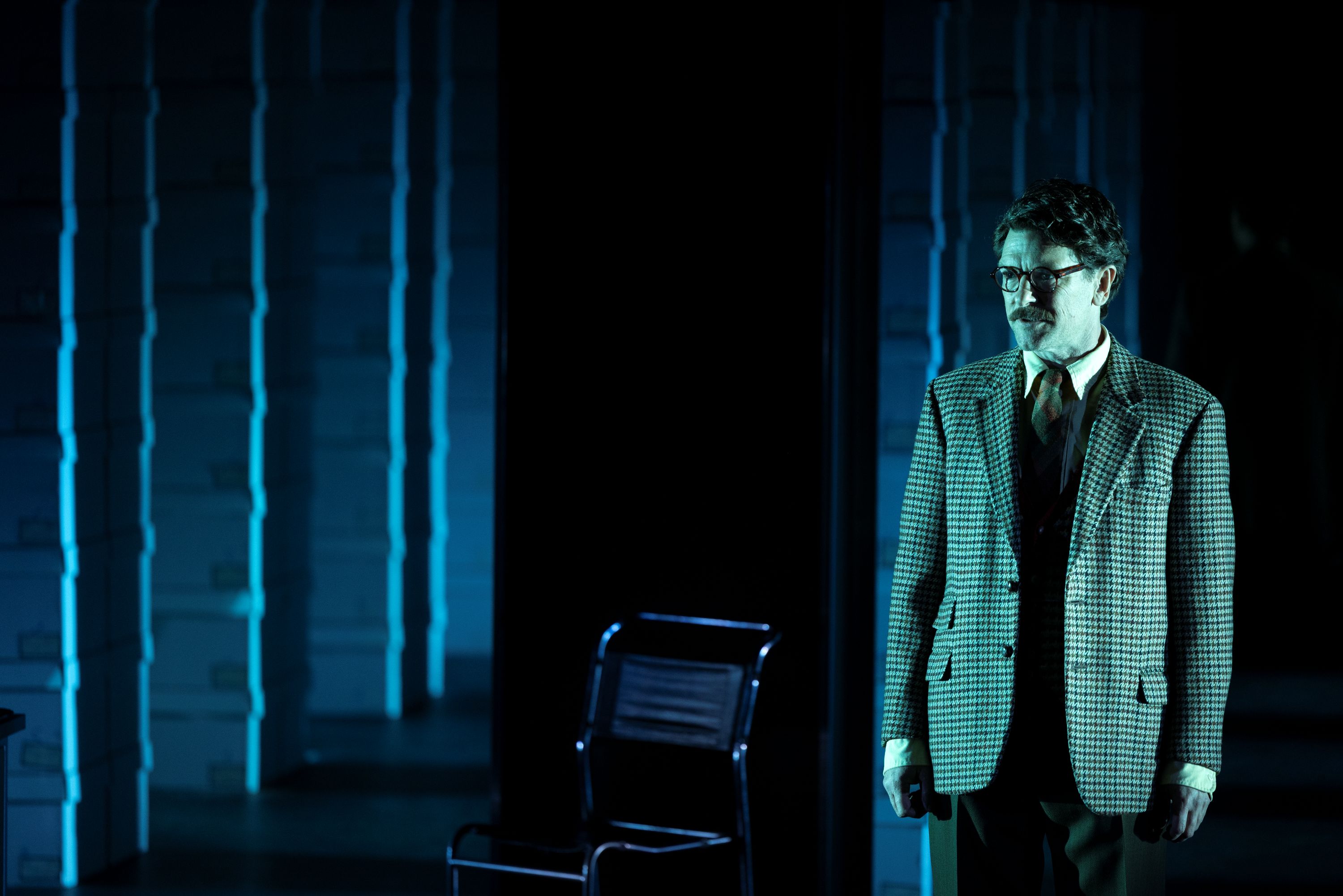 On stage, Dan Spielman, 47, in glasses and a tweed suit, stands, looking concerned, under blue light, by an office chair.