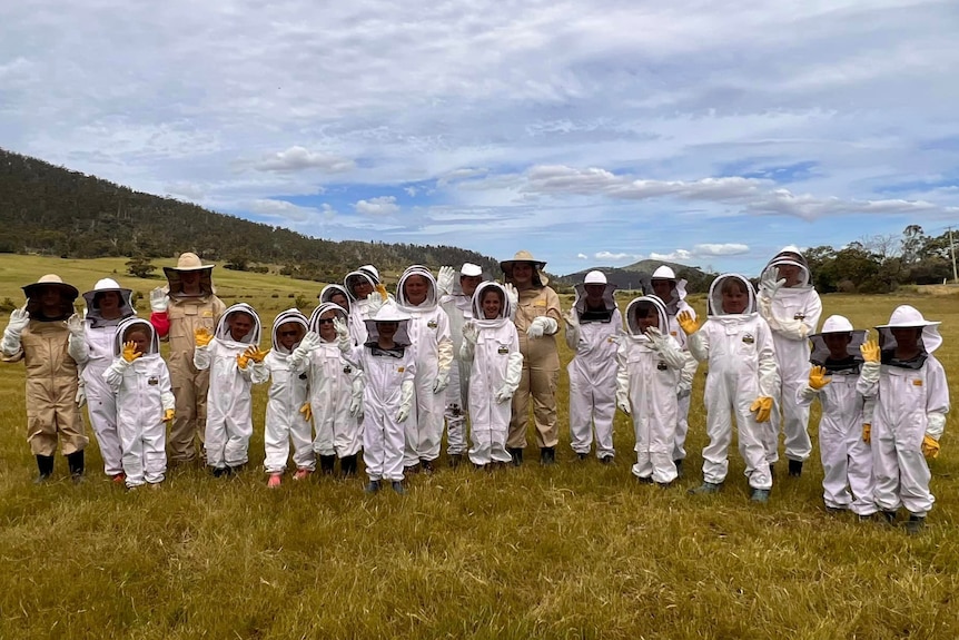 A group of beekeepers stand waving in a flat paddock with hills in background. Some are children and others are adults.