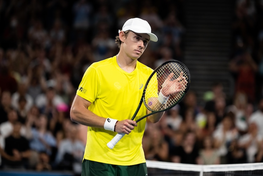 A young man in a yellow shirt and white cap with a tennis racquet in front of a crowd.