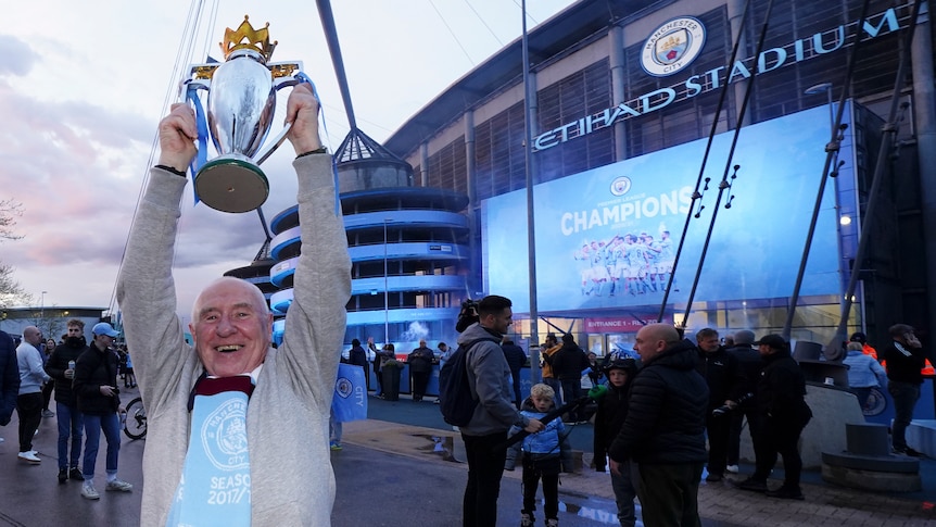 A grinning fan wearing a light blue shirt holds a replica cup above his head in front of a sign saying Champions.