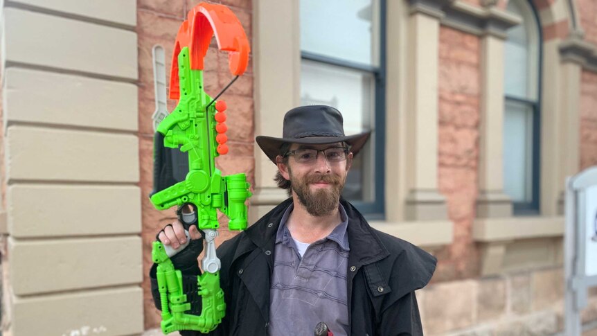 A man in a brown hat and coat with a sword at his belt holds a nerf cross bow aloft