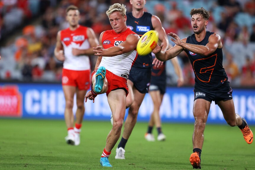 A blond-haired Sydney Swans forward snaps the ball towards goal with his right foot as a Giants defender tries to smother.