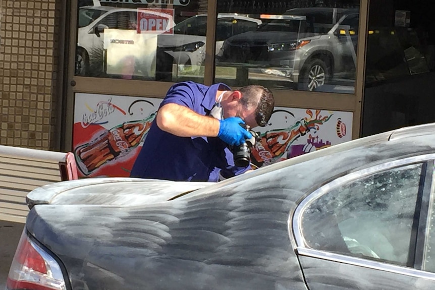 A forensic officer photographs a car dusted for fingerprints outside a shop.