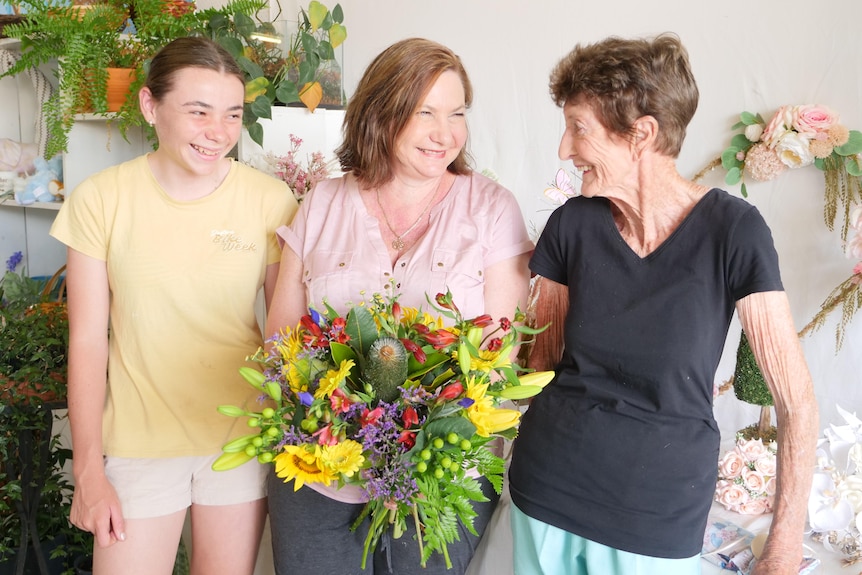 A woman stands with her adult daughter and granddaughter, holding a bouquet of flowers.