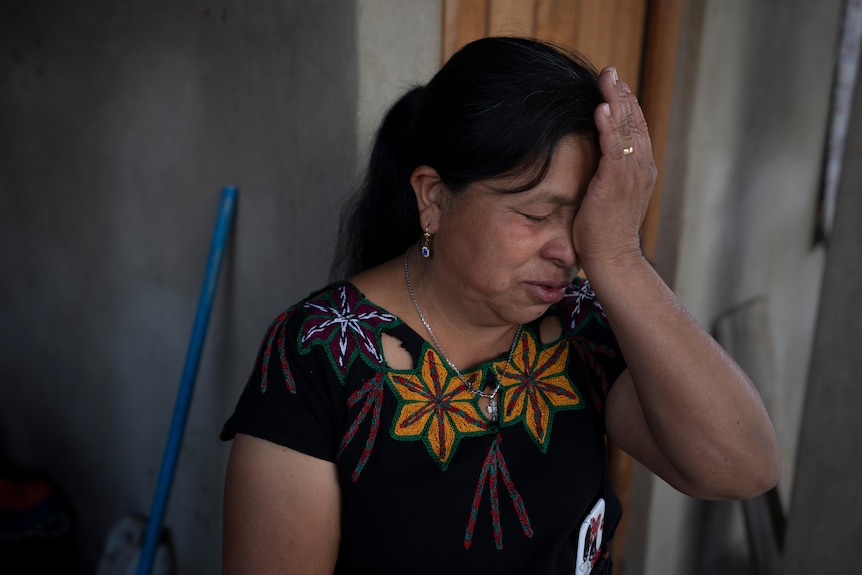 A Guatemalan woman reacts with eyes closed and hand against her forehead.