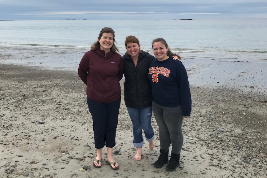 Three women in warm clothing standing on mild swamp-like, rock and weedy beach with blue ocean, overcast clouds in background