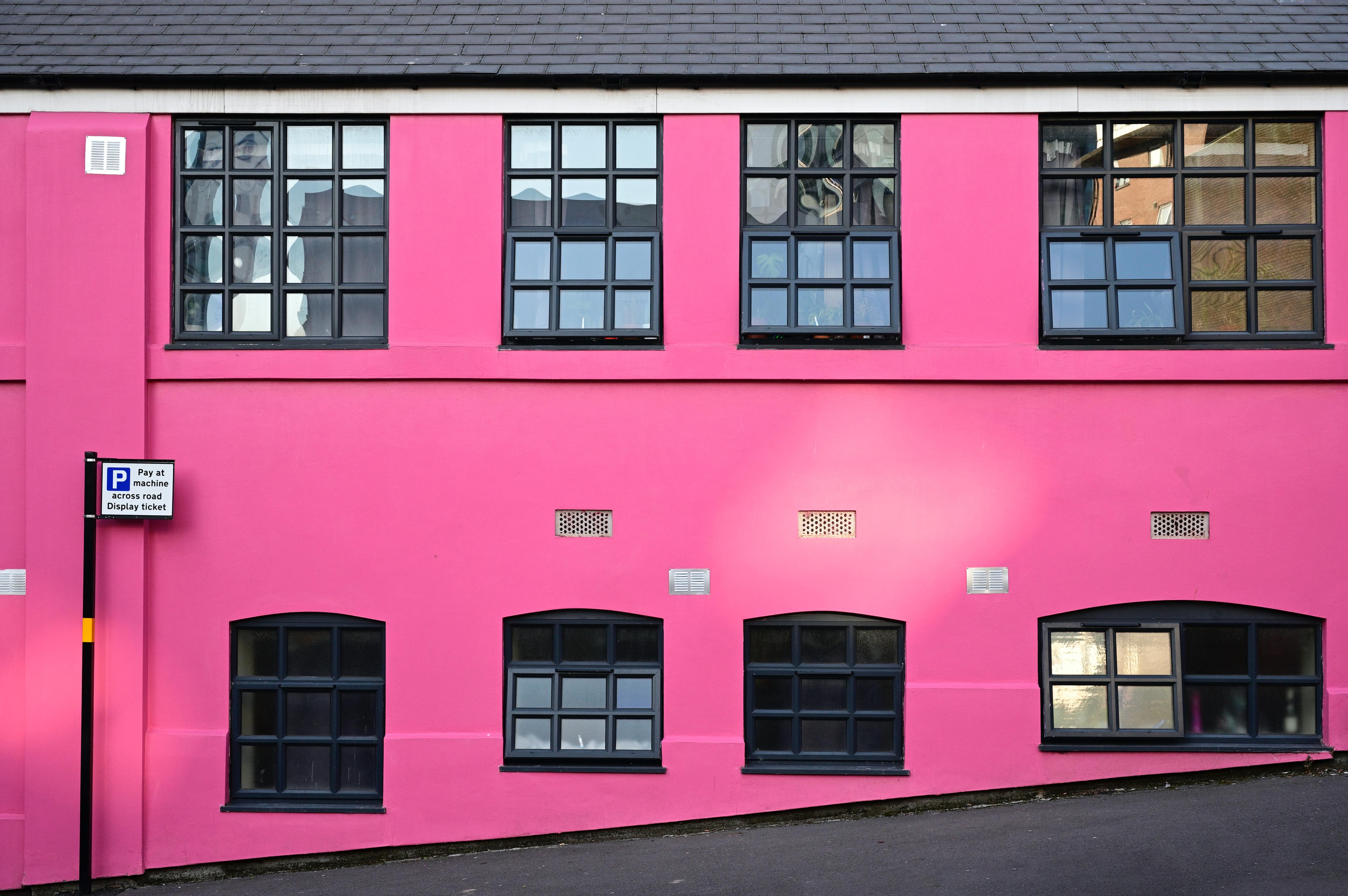 A bright pink building fascade with row of black-framed windows at top and bottom. Small 'P' sign stands at far left.