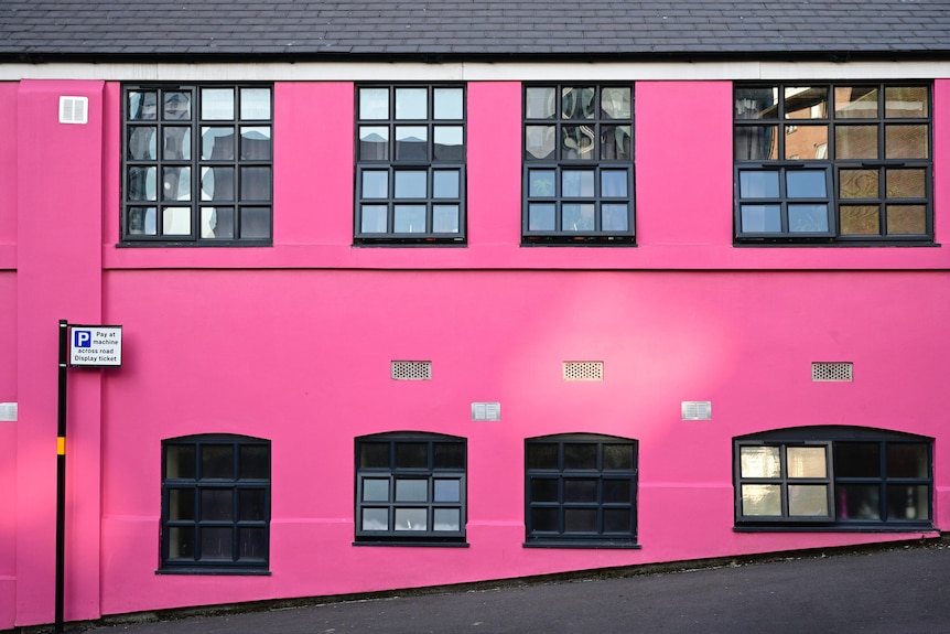 A bright pink building fascade with row of black-framed windows at top and bottom. Small 'P' sign stands at far left.