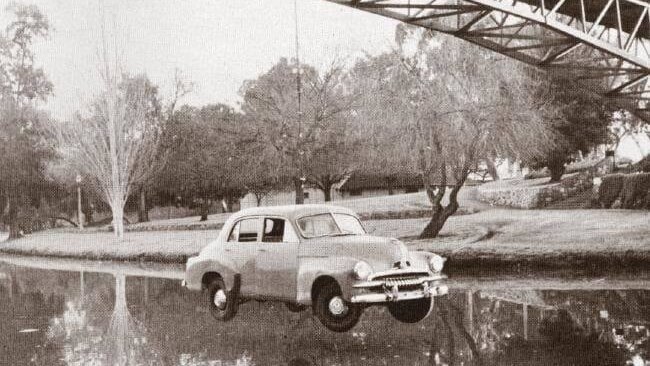 black and white image of men hanging an fj holden from a footbridge over a river 
