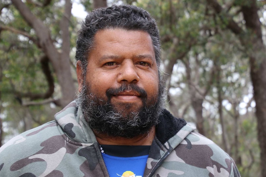 A head and shoulders profile shot of a middle aged Indigenous man with a beard and wearing a camo hoodie.