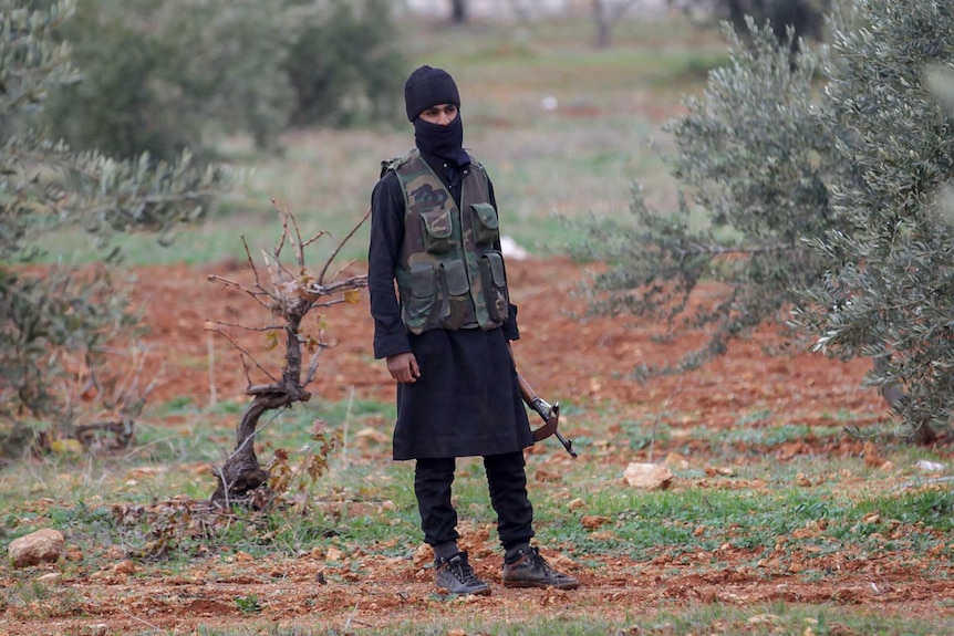 A man in an army vest and balaclava stands in a clearing in a field holding a gun