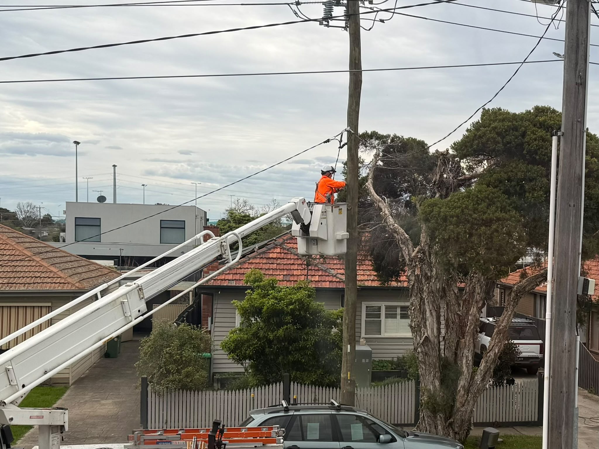 An electrician in orange hi-vis on a crame working on a power pole on a residential street.