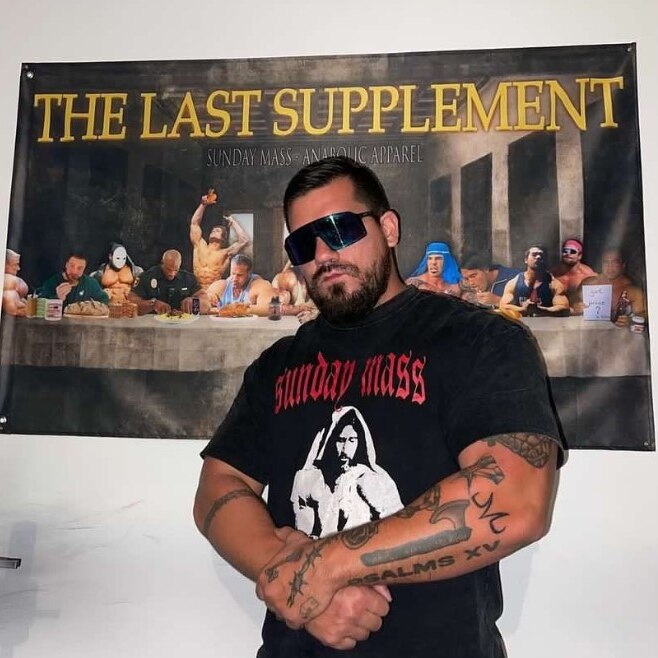 A picture of a muscular man with dark hair, fair skin, tattoos and sunglasses in front of a poster saying "the last supplement".