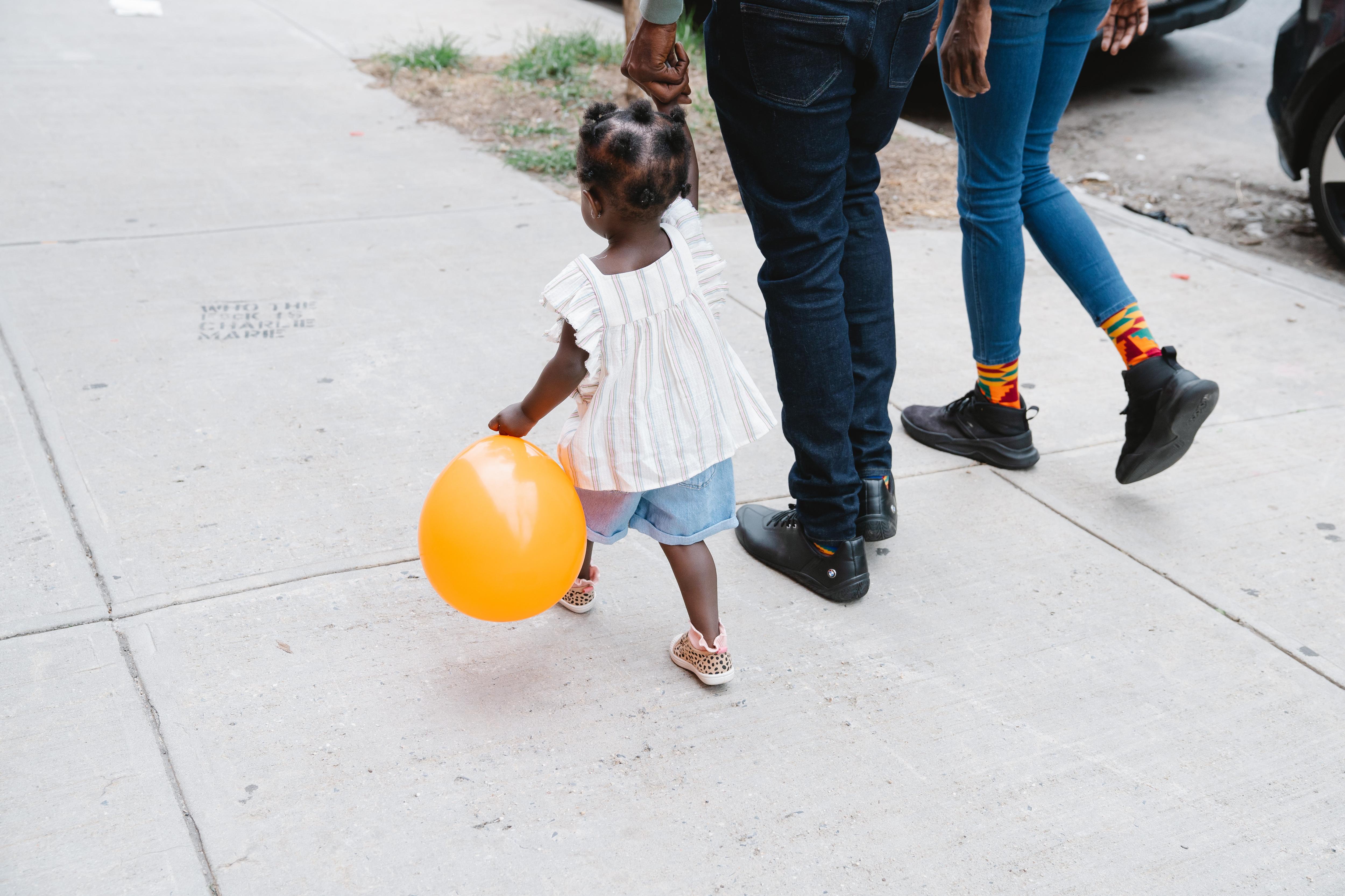 A little girl holding a yellow balloon and her parent's hand as they walk on a grey footpath.