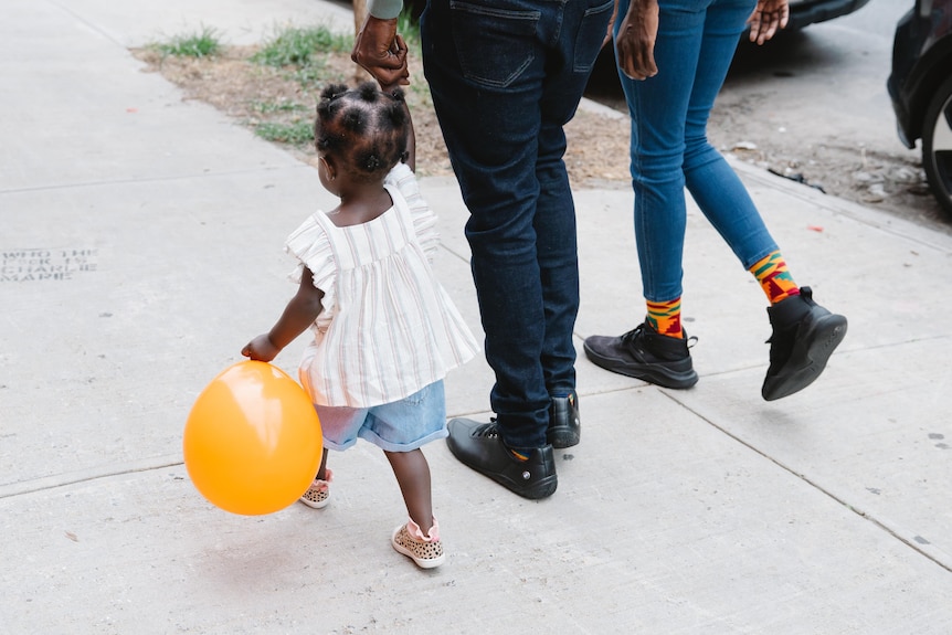 A little girl holding a yellow balloon and her parent's hand as they walk on a grey footpath.