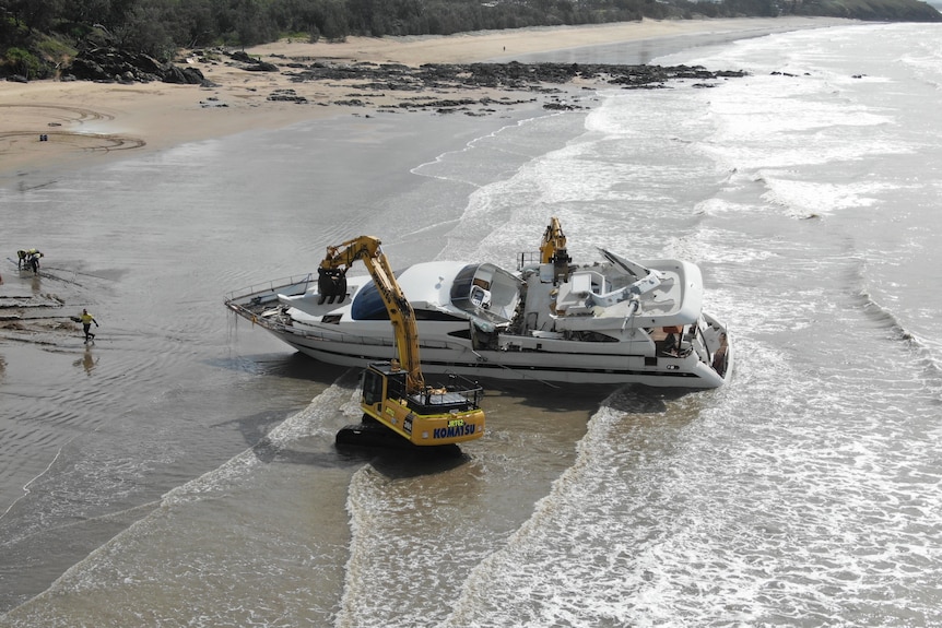 A digger pulls apart a yacht sitting in shallow waters at a beach